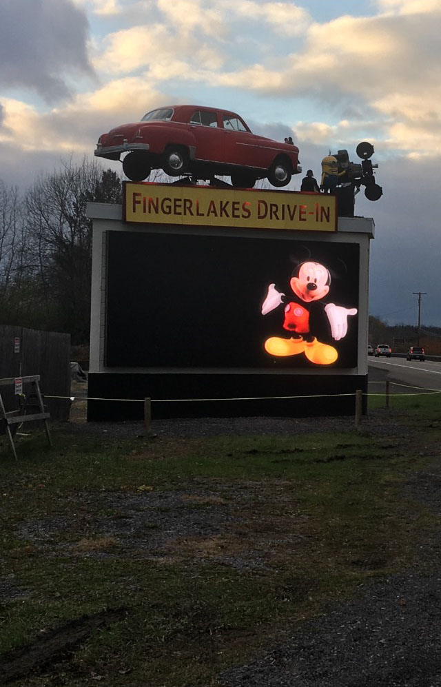 The Finger Lakes Drive-In marquee lit at dusk — a classic car and projector sculpture crowning the LED sign