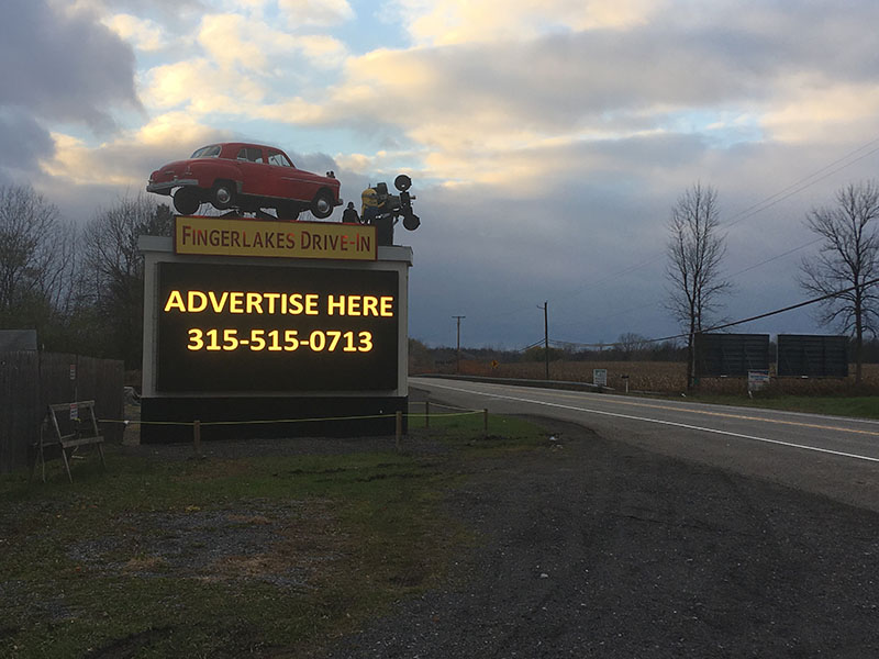 The Finger Lakes Drive-In marquee sign, topped with a classic car and film projector, against a dusk sky along Routes 5 & 20 in Aurelius, NY